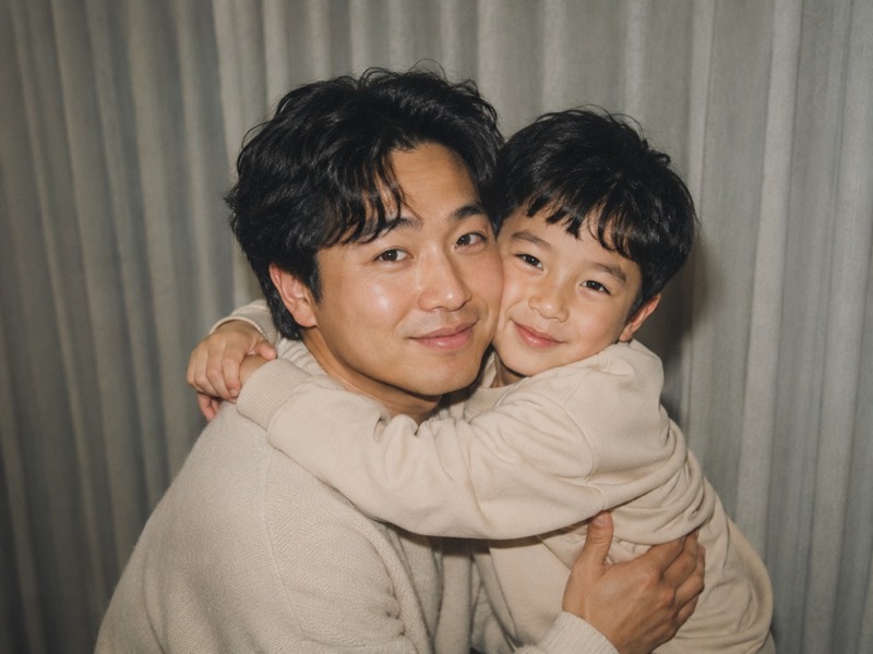 A man holding his younger self, both smiling, in front of pale curtains.